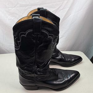 K&ouml;nnte beinhalten: Ein Paar schwarze Cowboystiefel aus Leder mit spitzer Zehenpartie, einem Blockabsatz und dekorativen, farblich abgestimmten Ziern&auml;hten am Schaft. Die Stiefel verf&uuml;gen &uuml;ber Anziehschlaufen am oberen Rand und ein glattes, poliertes Oberleder.