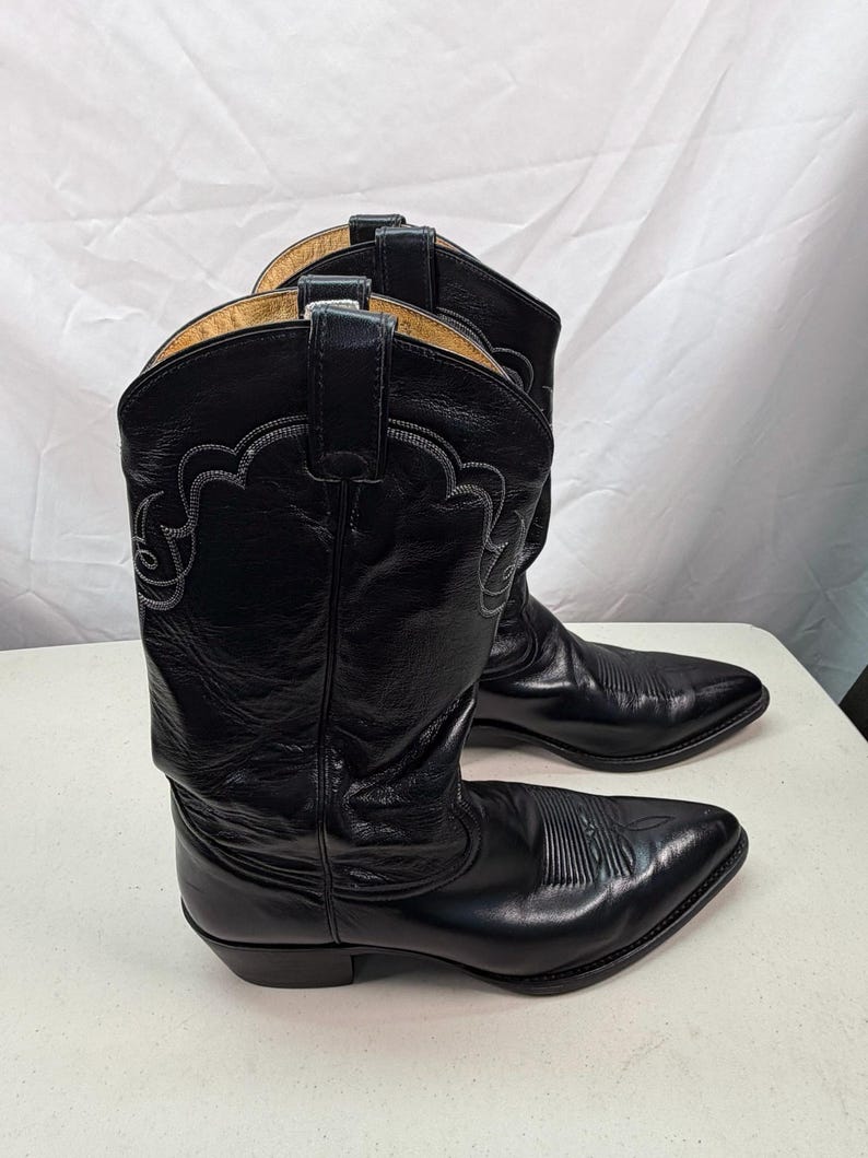 K&ouml;nnte beinhalten: Ein Paar schwarze Cowboystiefel aus Leder mit spitzer Zehenpartie, einem Blockabsatz und dekorativen, farblich abgestimmten Ziern&auml;hten am Schaft. Die Stiefel verf&uuml;gen &uuml;ber Anziehschlaufen am oberen Rand und ein glattes, poliertes Oberleder.