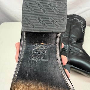 K&ouml;nnte beinhalten: Ein schwarzer Cowboystiefel aus Leder mit Absatz und einer strukturierten Gummisohle, die den Markennamen und das Sternlogo von Tony Lama tr&auml;gt. Die Sohle ist mit einem Echtleder-Stempel und der Tony Lama-Signatur versehen und weist Abnutzungen auf.