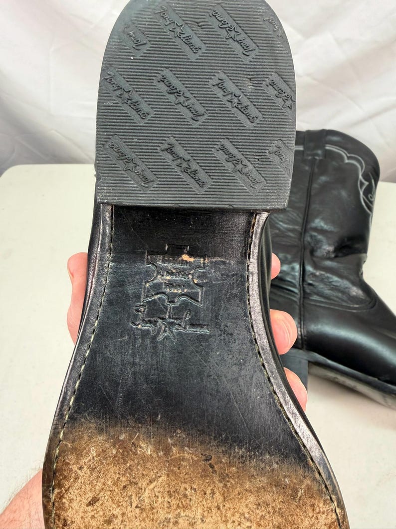 K&ouml;nnte beinhalten: Ein schwarzer Cowboystiefel aus Leder mit Absatz und einer strukturierten Gummisohle, die den Markennamen und das Sternlogo von Tony Lama tr&auml;gt. Die Sohle ist mit einem Echtleder-Stempel und der Tony Lama-Signatur versehen und weist Abnutzungen auf.