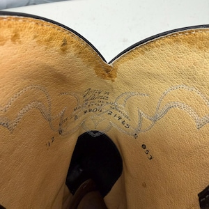 K&ouml;nnte beinhalten: Das Innere eines schwarzen Cowboystiefels aus Leder mit beigefarbenem Lederfutter. Der aufgedruckte Text enth&auml;lt 'Justin', 'MADE IN MEXICO', 'STYLE 9804', '21965' und 'SIZE D'. Dekorative Ziern&auml;hte sind entlang des oberen Schaftfutters sichtbar.