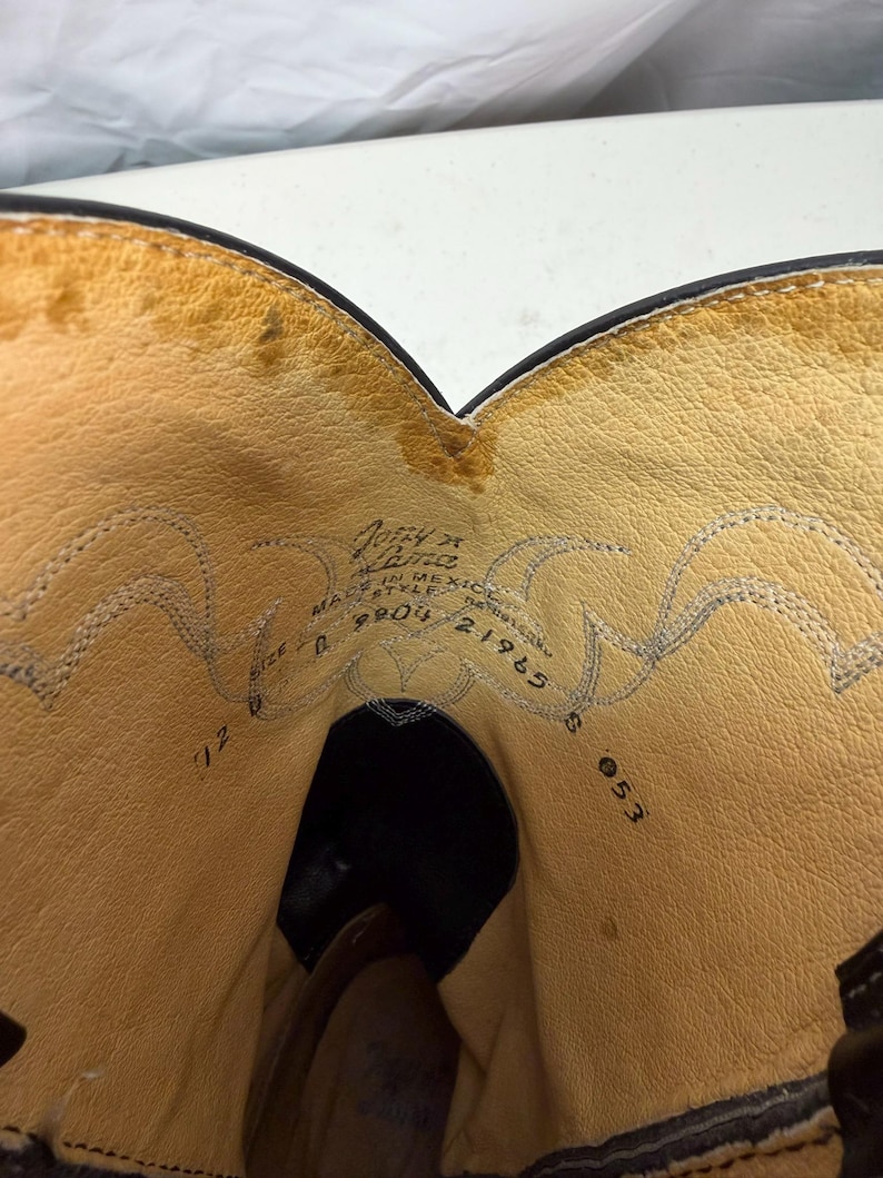 K&ouml;nnte beinhalten: Das Innere eines schwarzen Cowboystiefels aus Leder mit beigefarbenem Lederfutter. Der aufgedruckte Text enth&auml;lt 'Justin', 'MADE IN MEXICO', 'STYLE 9804', '21965' und 'SIZE D'. Dekorative Ziern&auml;hte sind entlang des oberen Schaftfutters sichtbar.