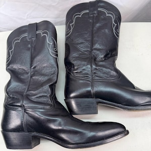 K&ouml;nnte beinhalten: Ein Paar schwarze Leder-Cowboystiefel mit wadenhohem Schaft, spitzer Zehenpartie und Blockabsatz. Der obere Schaft jedes Stiefels ist entlang der Oberkante mit dezenten, hellen, bogenf&ouml;rmigen Ziern&auml;hten versehen.
