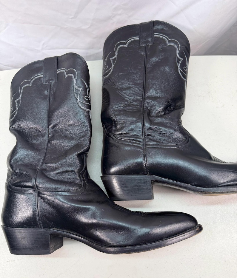 K&ouml;nnte beinhalten: Ein Paar schwarze Leder-Cowboystiefel mit wadenhohem Schaft, spitzer Zehenpartie und Blockabsatz. Der obere Schaft jedes Stiefels ist entlang der Oberkante mit dezenten, hellen, bogenf&ouml;rmigen Ziern&auml;hten versehen.