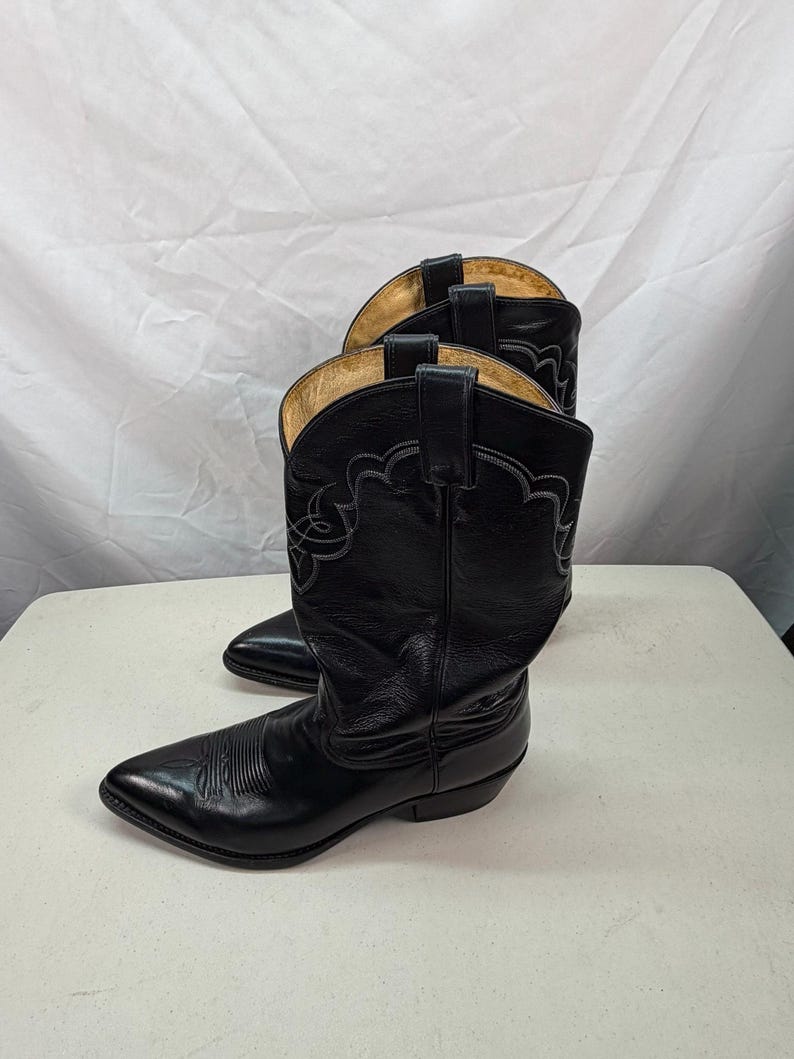 K&ouml;nnte beinhalten: Ein Paar schwarze Cowboystiefel aus Leder mit spitzer Zehenpartie, einem Blockabsatz und dekorativen grauen Ziern&auml;hten am Schaft. Die Stiefel verf&uuml;gen &uuml;ber Anziehschlaufen am oberen Rand und ein beiges Innenfutter in klassischer Western-Optik.