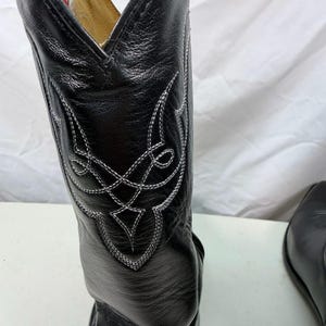 K&ouml;nnte beinhalten: Ein schwarzer Cowboystiefel aus Leder mit einem hohen Schaft und aufwendigen wei&szlig;en Ziern&auml;hten in einem geometrischen Muster. Der Stiefel hat eine spitz zulaufende Kappe und ein sichtbares Innenetikett mit rotem und blauem Text am Schaftrand.