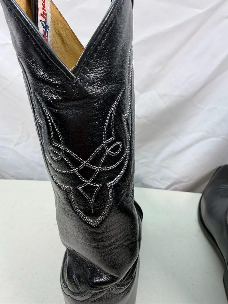 K&ouml;nnte beinhalten: Ein schwarzer Cowboystiefel aus Leder mit einem hohen Schaft und aufwendigen wei&szlig;en Ziern&auml;hten in einem geometrischen Muster. Der Stiefel hat eine spitz zulaufende Kappe und ein sichtbares Innenetikett mit rotem und blauem Text am Schaftrand.