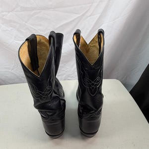 K&ouml;nnte beinhalten: Ein Paar schwarze Leder-Cowboystiefel mit hohem Schaft, V-f&ouml;rmigem Ausschnitt, Anziehschlaufen und dekorativen wei&szlig;en Ziern&auml;hten im oberen Bereich. Das Innere der Stiefel ist mit einem hellbraunen Material ausgekleidet.