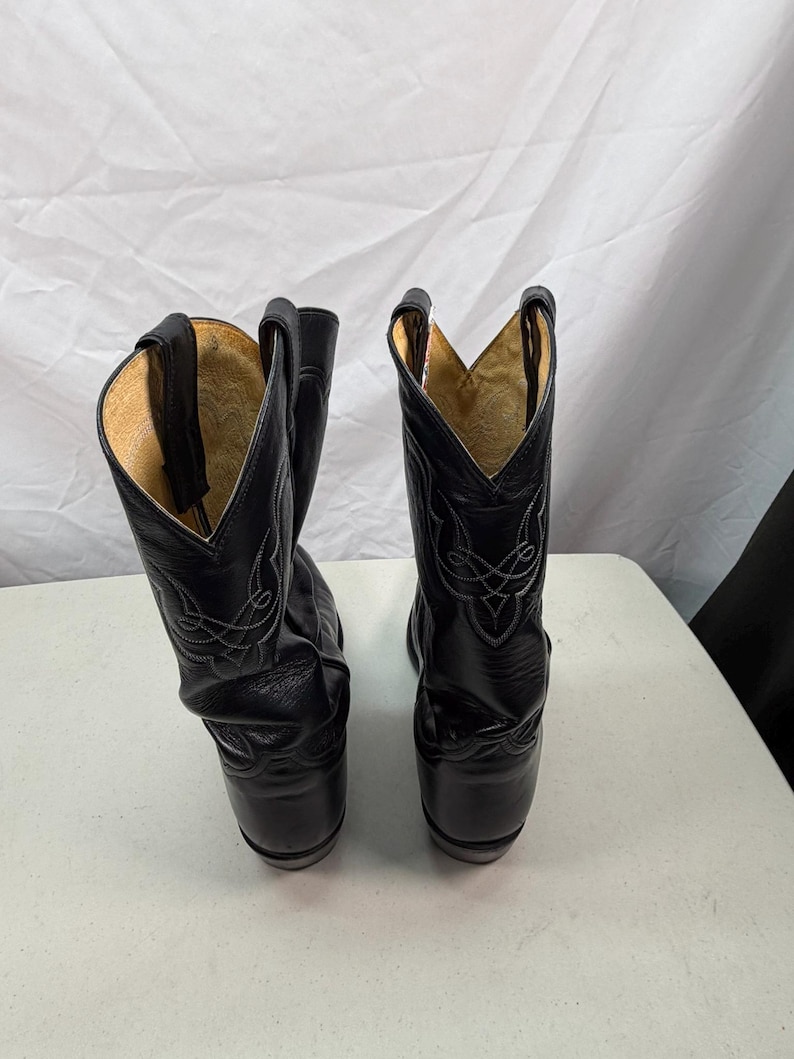 K&ouml;nnte beinhalten: Ein Paar schwarze Leder-Cowboystiefel mit hohem Schaft, V-f&ouml;rmigem Ausschnitt, Anziehschlaufen und dekorativen wei&szlig;en Ziern&auml;hten im oberen Bereich. Das Innere der Stiefel ist mit einem hellbraunen Material ausgekleidet.