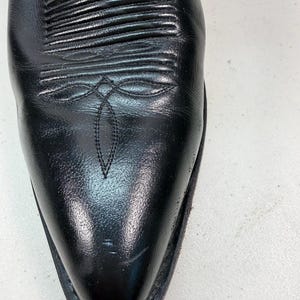 K&ouml;nnte beinhalten: Ein schwarzer Cowboystiefel aus Leder mit spitzer Zehenpartie und einem dekorativen Stickmuster auf dem Vorderblatt. Der Stiefel hat eine glatte, polierte Oberfl&auml;che mit horizontalen Rippendetails am Spann und einer dezenten Stickerei.