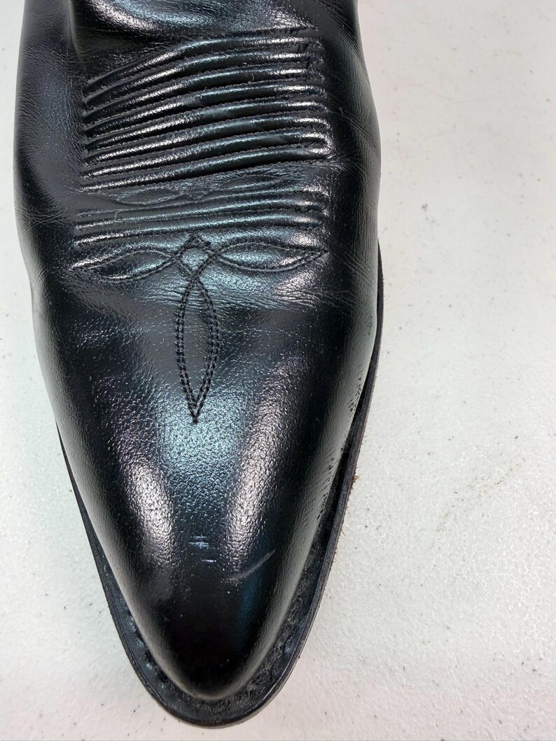 K&ouml;nnte beinhalten: Ein schwarzer Cowboystiefel aus Leder mit spitzer Zehenpartie und einem dekorativen Stickmuster auf dem Vorderblatt. Der Stiefel hat eine glatte, polierte Oberfl&auml;che mit horizontalen Rippendetails am Spann und einer dezenten Stickerei.