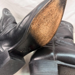 K&ouml;nnte beinhalten: Ein Paar schwarze Cowboystiefel aus Leder mit spitzer Zehenpartie und Absatz. Die Sohle eines Stiefels ist sichtbar und zeigt eine abgenutzte, strukturierte hellbraune Lederoberfl&auml;che. Die schwarzen Abs&auml;tze haben ein gepr&auml;gtes Logo.