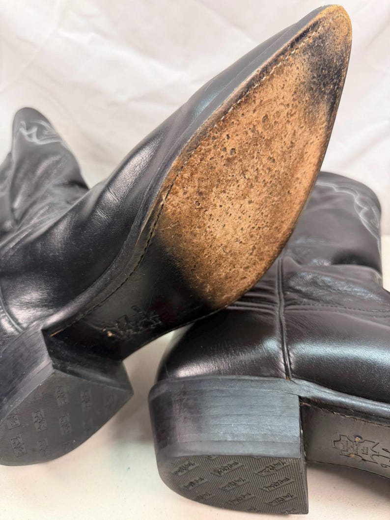 K&ouml;nnte beinhalten: Ein Paar schwarze Cowboystiefel aus Leder mit spitzer Zehenpartie und Absatz. Die Sohle eines Stiefels ist sichtbar und zeigt eine abgenutzte, strukturierte hellbraune Lederoberfl&auml;che. Die schwarzen Abs&auml;tze haben ein gepr&auml;gtes Logo.
