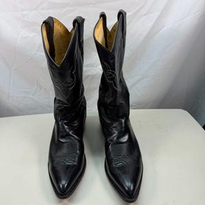 K&ouml;nnte beinhalten: Ein Paar schwarze Cowboystiefel aus Leder mit spitzer Zehenpartie, Absatz und dekorativen Ziern&auml;hten am Schaft und Vorderblatt. Das Innenfutter ist hellbraun und die Stiefel haben eine hohe, strukturierte Form mit Anziehschlaufen am oberen Rand.