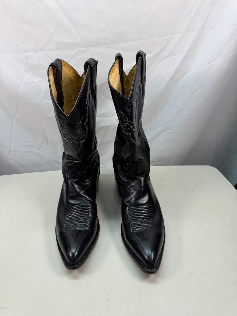 K&ouml;nnte beinhalten: Ein Paar schwarze Cowboystiefel aus Leder mit spitzer Zehenpartie, Absatz und dekorativen Ziern&auml;hten am Schaft und Vorderblatt. Das Innenfutter ist hellbraun und die Stiefel haben eine hohe, strukturierte Form mit Anziehschlaufen am oberen Rand.