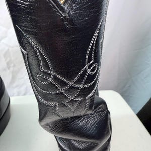 K&ouml;nnte beinhalten: Ein schwarzer Lederstiefel mit einem hohen Schaft und aufwendigen, wei&szlig;en Ziern&auml;hten. Das Leder weist eine strukturierte Narbung auf, und der Stiefel verf&uuml;gt &uuml;ber ein spitz zulaufendes Kragendesign an der oberen &Ouml;ffnung.
