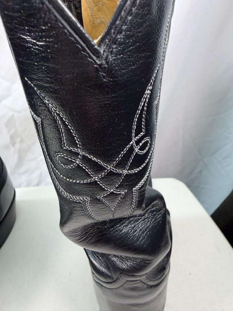 K&ouml;nnte beinhalten: Ein schwarzer Lederstiefel mit einem hohen Schaft und aufwendigen, wei&szlig;en Ziern&auml;hten. Das Leder weist eine strukturierte Narbung auf, und der Stiefel verf&uuml;gt &uuml;ber ein spitz zulaufendes Kragendesign an der oberen &Ouml;ffnung.