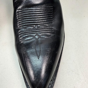 K&ouml;nnte beinhalten: Ein schwarzer Cowboystiefel aus Leder mit spitzer Zehenpartie und dekorativen Ziern&auml;hten auf dem Vorderblatt. Der Stiefel hat eine glatte Oberfl&auml;che mit sichtbaren nat&uuml;rlichen Falten und einen dunklen Sohlenrand im klassischen Western-Stil.