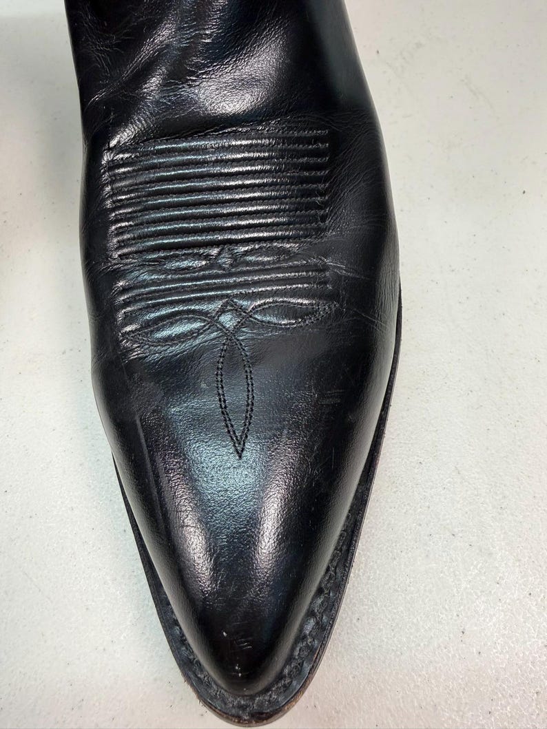 K&ouml;nnte beinhalten: Ein schwarzer Cowboystiefel aus Leder mit spitzer Zehenpartie und dekorativen Ziern&auml;hten auf dem Vorderblatt. Der Stiefel hat eine glatte Oberfl&auml;che mit sichtbaren nat&uuml;rlichen Falten und einen dunklen Sohlenrand im klassischen Western-Stil.