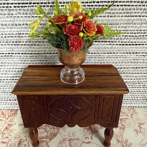 Peut inclure: Une petite table en bois brun foncé avec des détails sculptés. Un vase cuivré rempli de fleurs rouges et jaunes est posé dessus, créant une présentation décorative miniature.