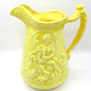 May include: A yellow ceramic pitcher with a detailed handle and spout. The pitcher features a raised relief design of classical figures. The pitcher is a light yellow color and has a glossy finish. A decorative piece for the home.