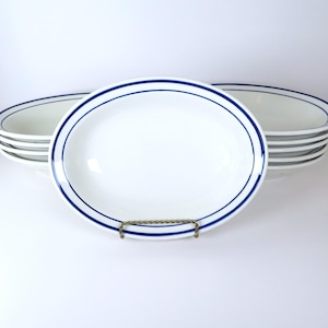May include: A collection of white, oval serving dishes with a double blue line border. The dishes are stacked on a white surface. One dish is in the foreground, supported by a gold-coloured wire stand.