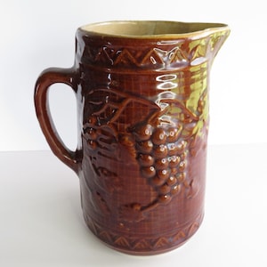 May include: A brown glazed ceramic jug featuring a raised relief pattern of grape clusters and leaves on a grid background. The vessel has a sturdy handle, a wide spout, and a decorative geometric triangle border along the top and bottom rims.