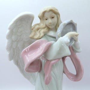 May include: A porcelain figurine of an angel with long blonde hair and large white wings. The angel wears a pale green robe with a flowing pink sash draped across the front and holds a small, light blue circular object in both hands.