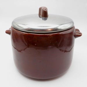 May include: A dark brown ceramic bean pot with a glossy finish, featuring two small side handles and a removable polished stainless steel lid with a central brown knob. The pot has a cylindrical body with a slightly rounded base and smooth surface.