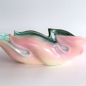 May include: An elongated ceramic bowl featuring a flowing, organic shape with a glossy finish. The exterior is a soft pastel pink that transitions into a creamy yellow base, while the interior displays a deep, contrasting teal green glaze.