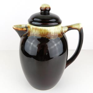 May include: A vintage brown ceramic coffee pot with a lid. The pot has a glossy finish with a two-tone design, featuring a dark brown base and a lighter brown and tan band near the top. It has a curved handle and a spout for pouring.