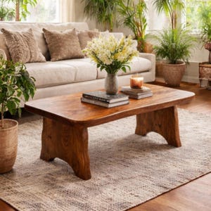 May include: A solid wood coffee table with a natural finish. The table has a rectangular top with rounded edges and sturdy, carved legs. A vase of white flowers, books, and a candle sit on the table. The table is in a living room setting.