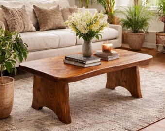 Puede incluir: Una mesa de centro de madera maciza con acabado natural. La mesa tiene una tapa rectangular con bordes redondeados y patas talladas. Un jarrón con flores blancas, libros y una vela están sobre la mesa. La mesa está en un salón.