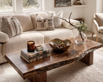 May include: A rectangular live-edge wooden coffee table with a dark brown finish and natural wood grain. The table features a thick slab top with rough, uneven edges supported by two sturdy block legs, holding a candle, books, and a bowl.