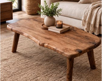 May include: A low-profile coffee table crafted from natural wood with a live edge, featuring a smooth, irregular oval surface and four sturdy, cylindrical legs. The wood grain is visible across the top, showcasing a warm, medium-brown tone.