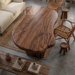 Puede incluir: Una gran mesa de centro de madera de forma org&aacute;nica con un rico acabado marr&oacute;n oscuro. La mesa est&aacute; sostenida por una base s&oacute;lida y se asienta sobre una alfombra tejida. La habitaci&oacute;n tambi&eacute;n cuenta con sillas de mimbre, peque&ntilde;os taburetes de madera y un sof&aacute;.