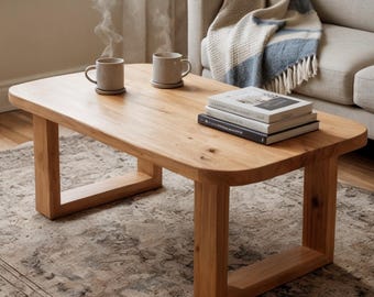May include: A light brown wooden coffee table with rounded corners and U-shaped legs. Two beige ceramic mugs with rising steam sit on the surface alongside a stack of three books. The table features a natural wood grain finish and sturdy construction.