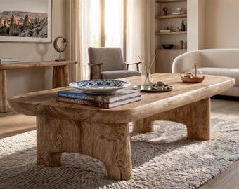 May include: A low-profile coffee table crafted from light-toned natural wood with a thick, organic slab top and sturdy, arched legs. The table surface holds a blue and white patterned ceramic bowl, two stacked books, and a glass reed diffuser.