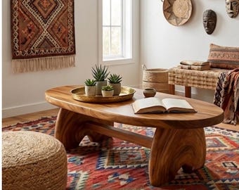 May include: An oval wooden coffee table with a smooth, natural grain finish and thick, curved legs. On the table sits a round, hammered gold metal tray holding four small potted succulents, alongside an open book with white pages.