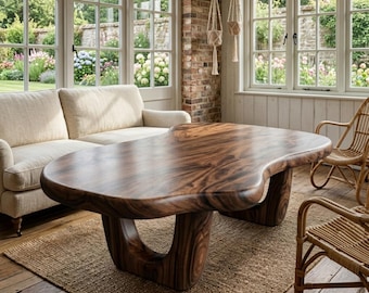 Könnte beinhalten: Ein großer, organisch geformter Couchtisch aus Holz mit einer satten, dunkelbraunen Oberfläche. Der Tisch hat ein einzigartiges, fließendes Design mit geschwungenen Kanten und stabilen, skulpturalen Beinen. Er steht auf einem gewebten Jute-Teppich in einem sonnigen Raum.