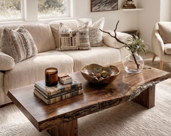Puede incluir: Una mesa de centro rectangular de madera con borde natural, acabado marrón oscuro y veta visible. La mesa presenta una superficie gruesa con bordes irregulares sostenida por dos patas de bloque robustas, con una vela, libros y un cuenco.