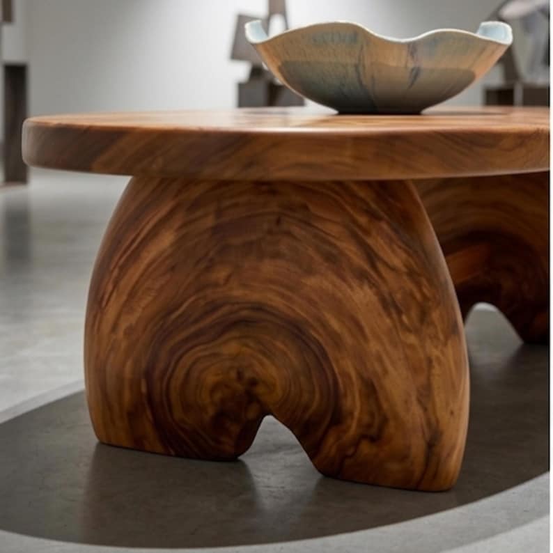 K&ouml;nnte beinhalten: Ein niedriger Couchtisch aus massivem Holz mit ausgepr&auml;gter nat&uuml;rlicher Maserung und dicken, bogenf&ouml;rmigen Beinen. Eine flache, breite Keramikschale mit gewelltem Rand und blau-wei&szlig;er Farbverlaufglasur steht auf der glatten, runden Tischplatte.