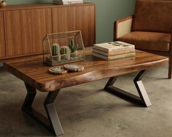 May include: A rectangular wooden coffee table with a live edge and black metal legs. The table has a natural wood grain finish and is decorated with a glass terrarium with cacti, coasters, and books. The table is in a living room setting.