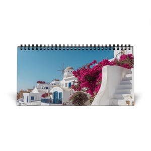 Könnte beinhalten: Ein Spiral-Schreibtischkalender mit einem Foto eines griechischen Dorfes. Das Bild zeigt weiße Gebäude, blaue Akzente und leuchtend rosa Blumen. Der Kalender ist geöffnet und zeigt die malerische Szene.