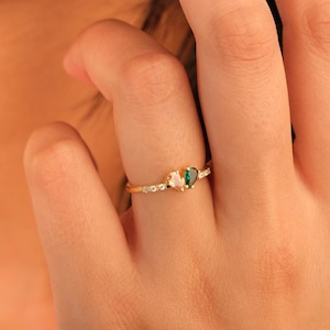 May include: A gold ring with a small emerald-green heart-shaped stone, a white stone, and small clear stones. The ring is on a finger. The ring's design is delicate and elegant. The hand is in the foreground.