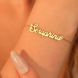 Op de afbeelding: Een goudkleurige armband met de naam "Seraphina" in cursief schrift. De fijne kettingarmband wordt om een pols gedragen, met de naam als middelpunt. De achtergrond is wazig, waardoor de sieraden worden benadrukt.