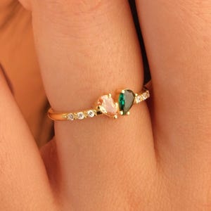 May include: A gold ring featuring a thin band set with small clear stones. The center of the ring displays two pear-shaped stones, one light pink and one emerald green, positioned side by side. The metal band has a polished finish and a delicate design.