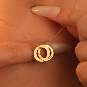 May include: A gold-toned necklace featuring two interlocking circular pendants. The pendants are engraved with the name MARGOT and the date 11.21.21. The necklace is suspended from a delicate gold-toned chain with a fine link design.