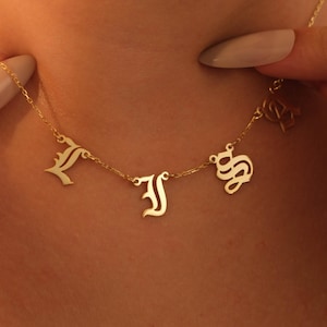 May include: A gold-toned necklace featuring a delicate chain with three distinct hanging letters in an ornate, gothic-style font. The letters are spaced out along the chain, which rests against the skin, showcasing a polished metallic finish.