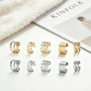 May include: A collection of gold and silver ear cuffs in various designs. Styles include double bands, leaf patterns, and textured surfaces. The jewelry is displayed on a white surface with a magazine in the background.